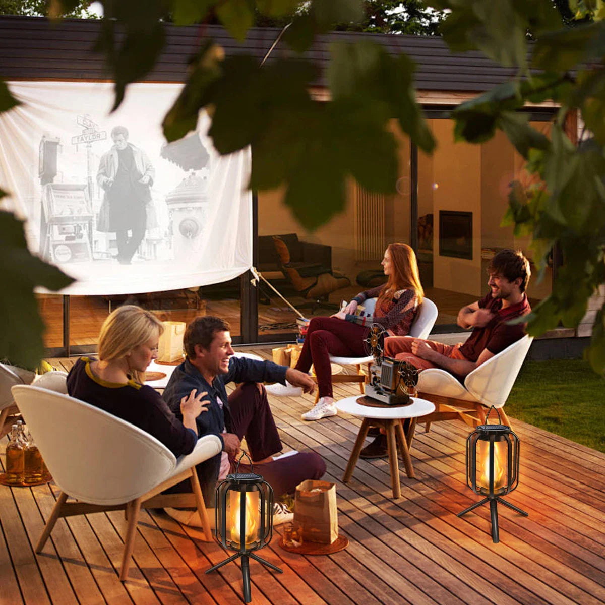 People watching a movie on a screen outdoors with lanterns on a wooden deck.